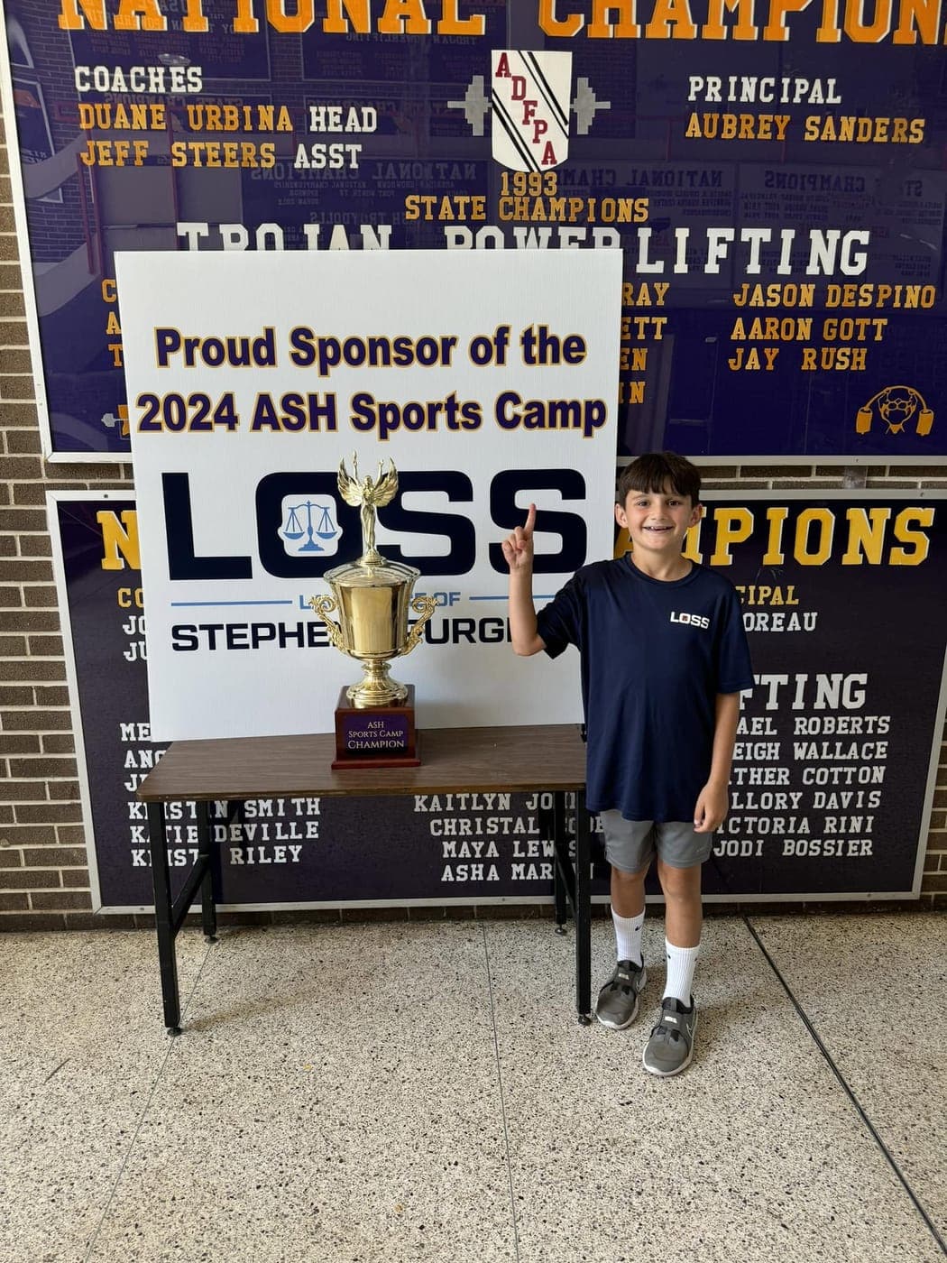 A camper beside the 2024 ASH Sports Camp champion trophy and the firm's sponsor sign